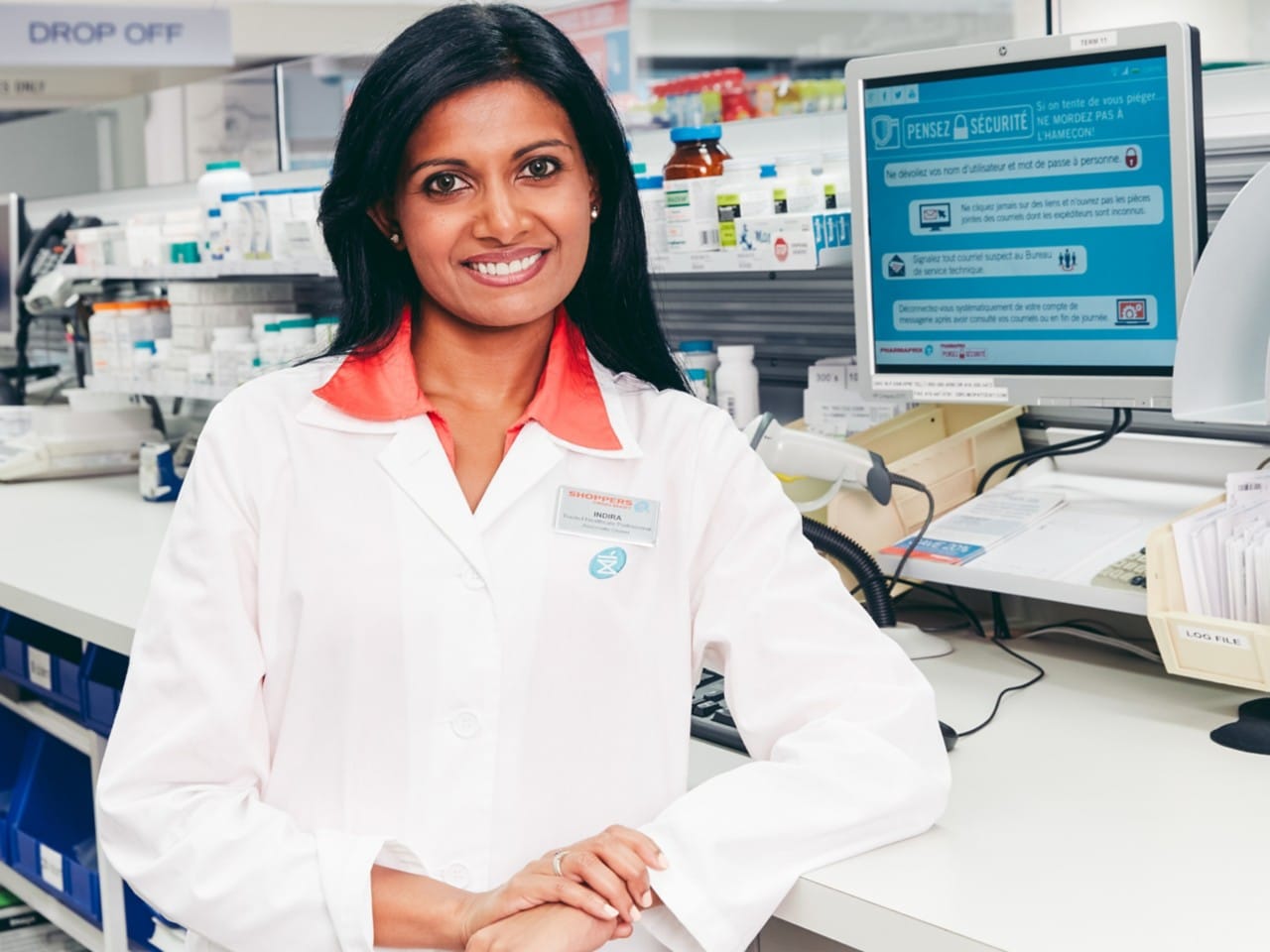 Pharmacist in white lab coat in front of a computer screen smiling.